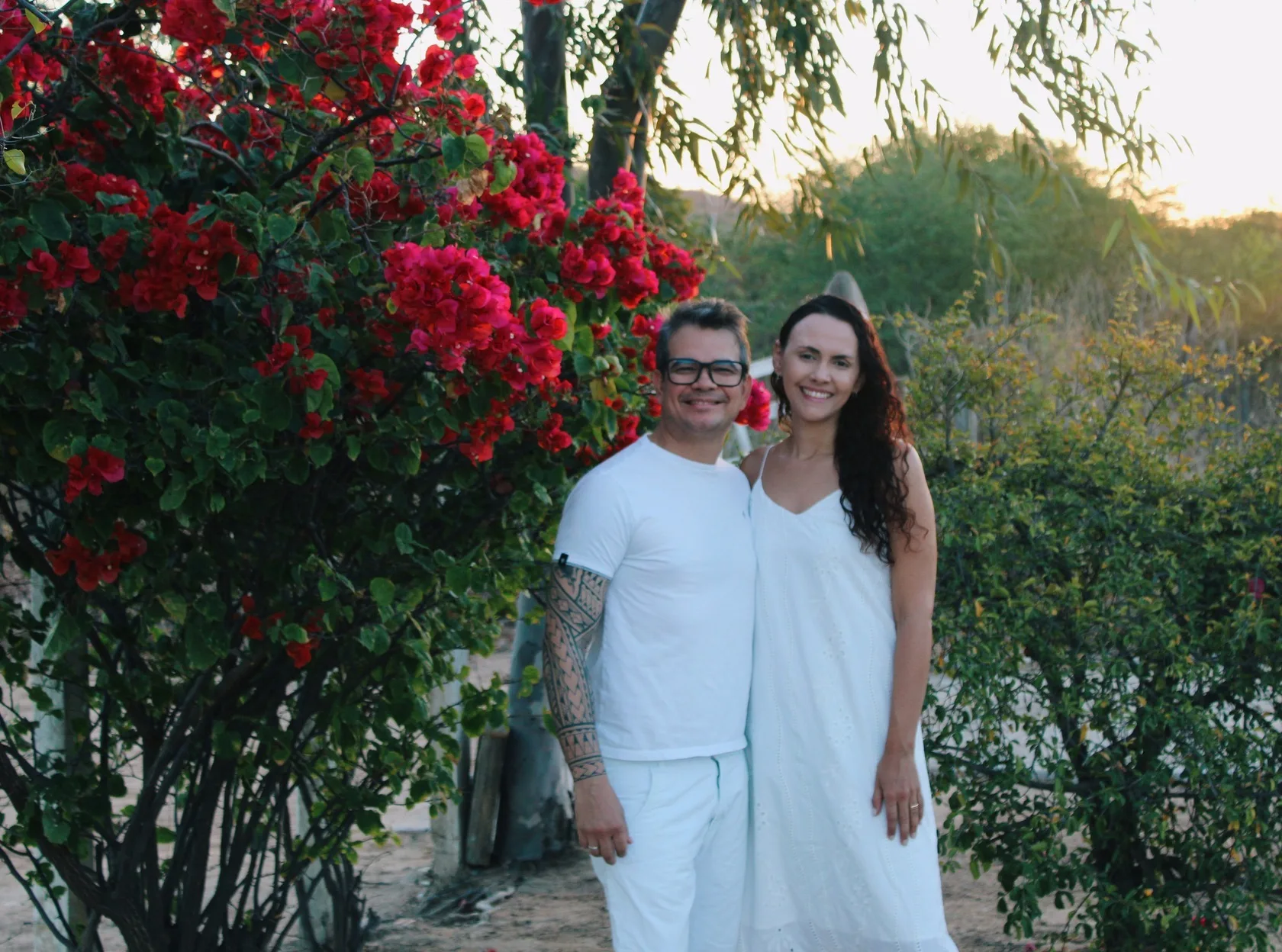 Casal de roupa branca sorrindo ao lado de um arbusto de flores vermelhas, ao entardecer.
