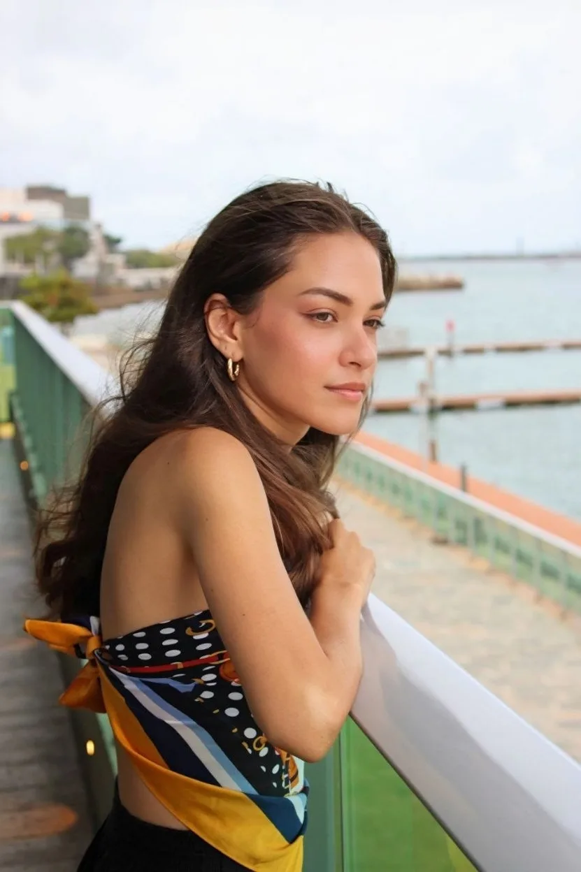 Jovem de perfil em uma passarela à beira-mar, de vestido estampado amarrado nas costas.