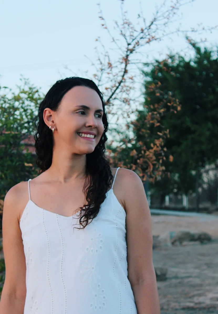 Mulher de vestido branco de alças sorrindo ao ar livre, com árvores ao fundo.