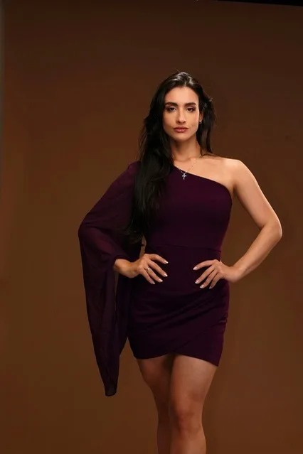 Mulher de vestido bordô de uma manga, com as mãos na cintura, em estúdio com fundo marrom.