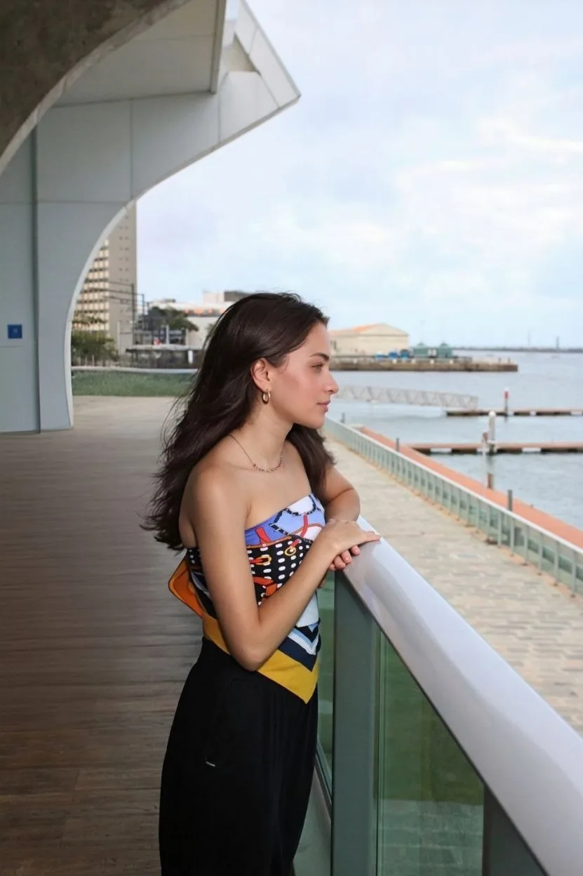 Jovem de vestido estampado e saia preta em uma passarela coberta à beira-mar.