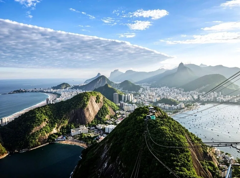 Vista aérea de montanhas cobertas de mata junto ao mar, com a cidade e a praia ao fundo.