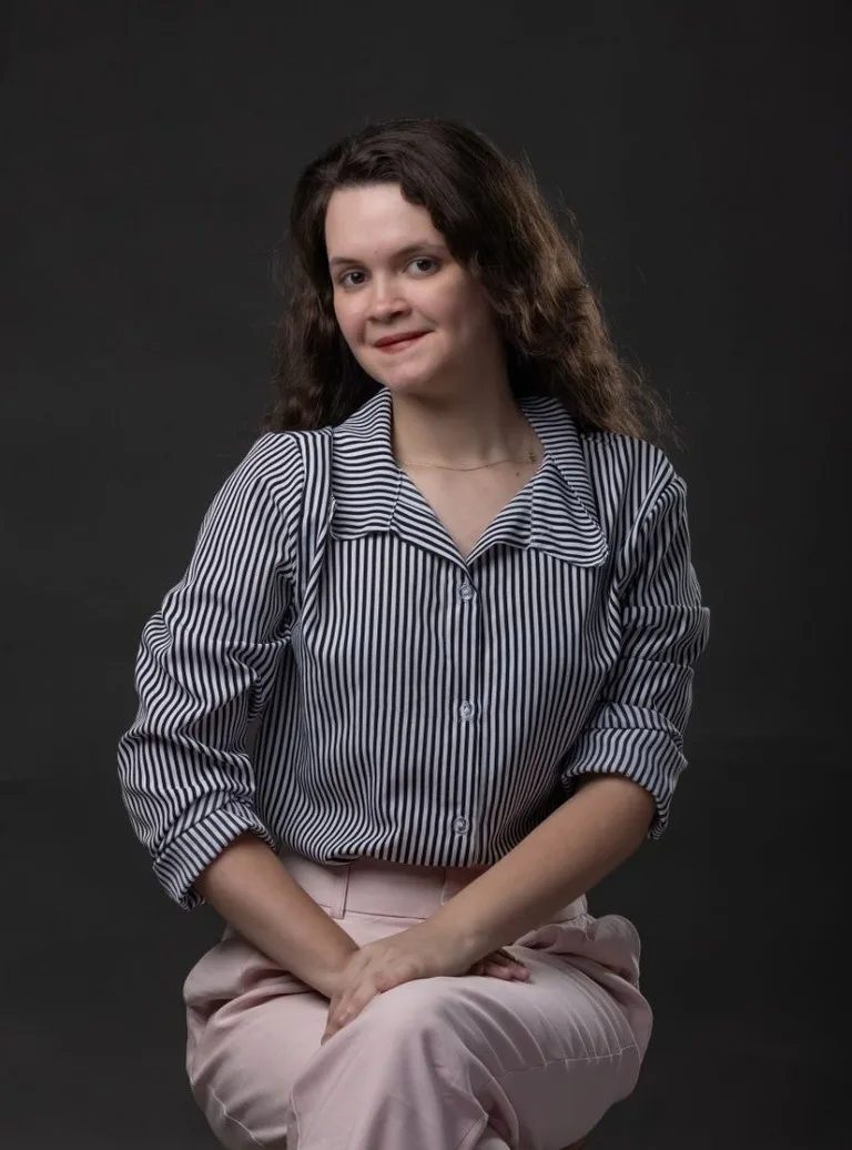 Luana Oliveira sentada em estúdio com fundo escuro, de camisa listrada e calça rosa-claro.