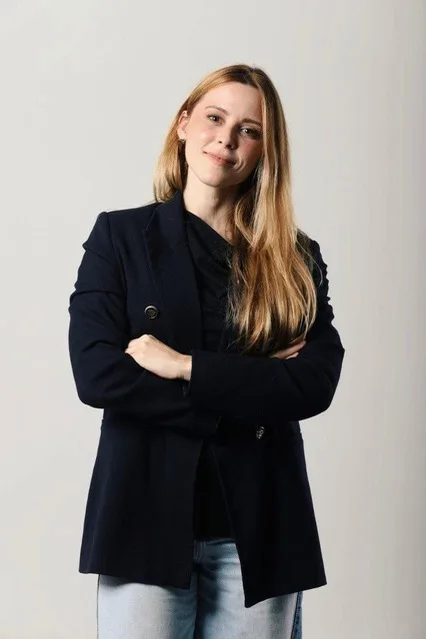 Mulher loira de blazer preto e braços cruzados, em estúdio com fundo claro.