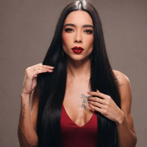 Mulher de cabelo preto longo, batom vermelho e vestido vermelho, em estúdio com fundo marrom-acinzentado.