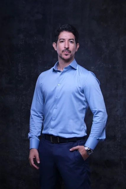 Homem de camisa azul-clara e calça escura, em estúdio com fundo azul-escuro.