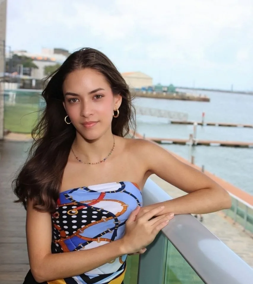 Jovem de cabelos longos e vestido estampado apoiada no guarda-corpo, olhando para a câmera, com o mar ao fundo.